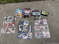 Lot displayed on concrete ground showing sports cards arranged in groups, unopened Hot Wheels Beatles Yellow Submarine car blister pack, Jeff Burton NASCAR car blister pack, Hangman handheld game, and Sudoku handheld game in packaging.