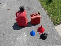 Photo showing two red gas cans and three funnels of different colors on concrete outdoors.
