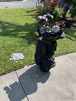 Outdoor photo of golf bag standing on concrete next to lawn, showing multiple golf clubs in the bag and a pile of golf balls plus wooden tees on the ground beside it.