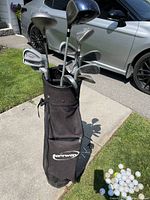Full view of black Hiway golf bag filled with various right-handed golf clubs and a pile of white golf balls and tees on the grass beside it