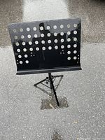 Front view of black metal adjustable music stand showing perforated sheet holder and tripod base on asphalt surface.