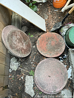 Seven round reddish-brown concrete patio stones arranged on dirt with surrounding outdoor objects.