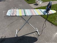 Image showing ironing board set up outdoors with iron placed on the metal rest and cord plugged in, highlighting condition and fabric pattern.