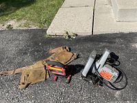 Photo showing a worn leather carpenter belt, an electric Skil circular saw, and a red trouble light on pavement.