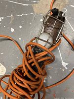 Close-up of handheld trouble light wrapped with an orange cord, metal cage for bulb visible. Shows signs of wear and use.