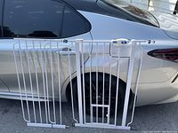 Photo showing white metal adjustable gate with vertical bars, including a small cat door, placed in front of car door for scale.