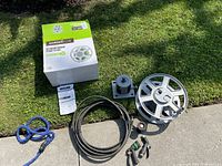 Overview of the lot showing the green and white Garden Club 60 ft roll-up water reel in box, black soaker hose, gray hose reel stand, and multiple hose sprayers on grass.