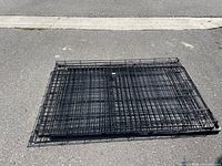 Photo of folded black metal wire dog crate placed flat on asphalt, showing the crate folded to a compact shape.