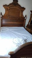 Front view of East Lake style antique wooden double bed with elaborate carved headboard, showing mattress and bedding in place. Wall and partial furniture on side visible.