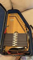 Wooden Appalachian autoharp in black hard case with red and black strap attached to the instrument.