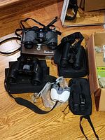 Overview of all five binoculars laid out on wooden floor showing various sizes and colors including textured gray Bushnell Insta-Focus Explorer and white compact binoculars.