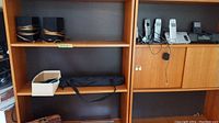 Two black and wooden Energy speakers and multiple cordless phones along with a tripod bag placed on wooden shelves.