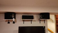 Photo showing both Bose 301V speakers mounted on black wall brackets on a white shelf above a TV.