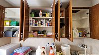 Open cabinets showing multiple shelves with various cleaning supplies, oils, sprays, detergents, insect repellents, cleaning brushes, scrubbers, batteries, light bulbs, and boxed items such as floodlights and matches.