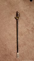 Full view of vintage decorative sword with black grip, brass hand guard, and scabbard lying on carpet.