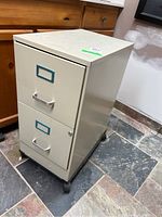 Full view of the beige two drawer metal filing cabinet on wheels placed on a tiled floor near wood cabinets.