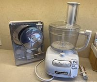 White KitchenAid food processor base with clear work bowl and feed tube next to a gray plastic case holding multiple stainless steel discs for slicing and shredding.