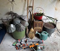 Wide shot showing metal garden lighting decor, plastic garbage can, broom, shovel, watering can, Gardena sprinklers and sprayers arranged on concrete floor against plaster wall.