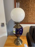 Full view of vintage table lamp with large white glass globe shade, blue ceramic base with gold floral design, metal bottom base and collar, and clear glass chimney on top.