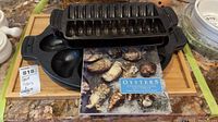 Two black cast iron Outset trays, one oyster-shaped with multiple oyster molds, and one shrimp-shaped with shrimp molds, along with an oyster guide and cookbook, all placed on a countertop.