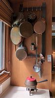 Copper pans, cake pans, metal ladles hanging on a vertical rack mounted on a wooden wall. Variety of pan sizes and shapes, including one small ladle and a metallic scoop.