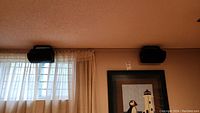 Two Polk Audio black speakers mounted on a wall above a window with curtains.