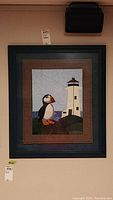 Full view of framed quilt showing puffin and lighthouse design inside dark frame on beige wall