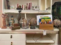 Wide shot showing various figurines including nude woman statue with container, a girl figurine in blue dress with teddy bear, tall bird figurine, and small copper pieces on counter. Also shows notecards and art supplies box.