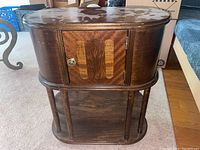 Front view of medium-sized brown wood smoker's table with oval top and decorated door with metal knob, standing on four cylindrical legs with open lower shelving