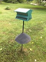 Full view of green metal bird feeder mounted on black metal post with large metal baffle, standing in grass outdoors.
