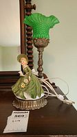 Front view of the lamp showing porcelain doll with green dress, metal base, and green glass shade