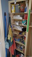Full view of shelving unit containing cleaning liquids, multiple white plastic hangers, and other cleaning supplies. Two flags hanging to the left with brooms and dustpan visible.