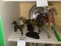 View of five horse figurines including brown ceramic foal, silver-tone metal horse, black glass horse, decorated black and yellow horse, and modern tan horse with rider. Pewter horse face plate at the back.