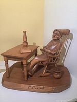 View of the entire wood carving showing man in rocking chair next to table with lamp and book on wooden base signed C Caron