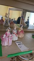 View of three Royal Doulton figurines displayed on a shelf in front of a mirror showing characters in historical dress.