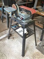 Front and side view of Delta 4 belt and 6 inch disc sander mounted on metal stand with red sanding belt and sanding disc visible. Instruction manual on lower shelf.