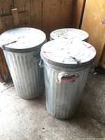 Photo shows three cylindrical metal trash cans with fitted lids arranged on a concrete floor inside a wooden structure. Two are larger and one smaller.