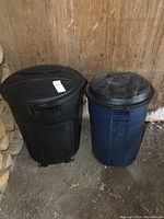 Pair of two Rubbermaid trash cans, black one with wheels on left, blue one without wheels on right, both with lids.