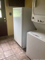 Freestanding white upright freezer next to stacked washer/dryer on tiled floor