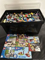Top-down view of black storage bin filled with assorted Lego pieces with visible building manuals in front
