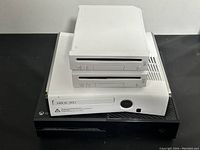 Two white Nintendo Wii consoles stacked on one Xbox 360 console beside a separate Xbox 360 console on black surface against white wall.