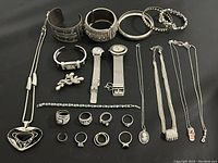 Full overview of assorted silver tone jewelry including watches, bracelets, rings, necklaces, and earrings laid flat on black background.