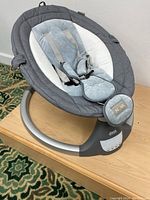 Full view of the Joie Nova baby rocker/soother placed on a wooden surface showing the whole frame, padded seat, and harness.