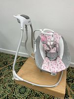 Right side view of the pink floral cushioned baby swing with gray padded sides, safety harness, and white support frame.