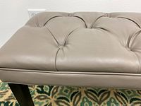 Close-up photo showing tufted cushion in greige faux leather with button details and part of a black wooden leg.