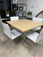 Square commercial dining table with hardwood top and polished stainless steel base, surrounded by four white chairs with wooden frame edges and polished stainless steel legs.