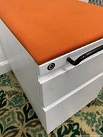 Close-up photo showing the cabinet's orange cushioned seat, white metal body with minor scratches, and black handle with key lock