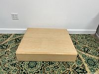Square wooden display stand on a patterned carpet, natural finish with visible wood grain.