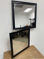 Two black framed square mirrors positioned one on the wall and one leaning against the wall on a wood surface. Shows overall design and size.