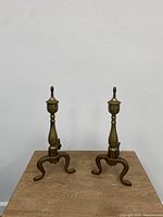 Front view of pair of bronze plated andirons with three-legged bases and turned finials, showing patina and wear
